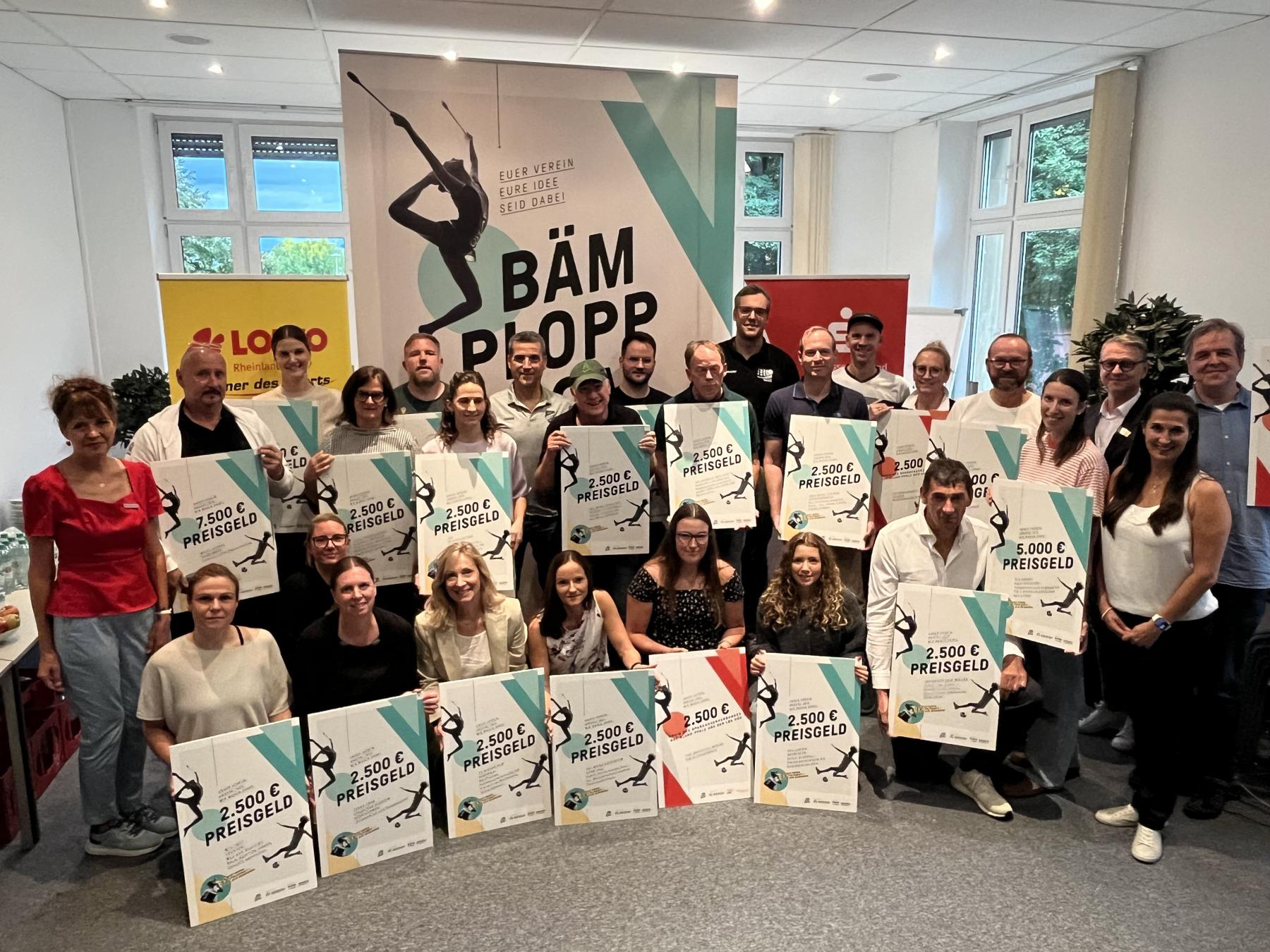 Gruppenbild in einem Raum mit siebenundzwanzig Personen mit Urkunden, in der Mitte ein Aufsteller: Bämploppboom mit einer Tänzerin der rhythmischen Sportgymnastik, im Hintergrund Aufsteller von Lotto und Sparkasse