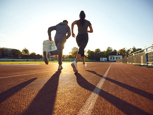 eine Frau und ein Mann in Sportkleidung laufen auf einer Tartanbahn im Gegenlicht