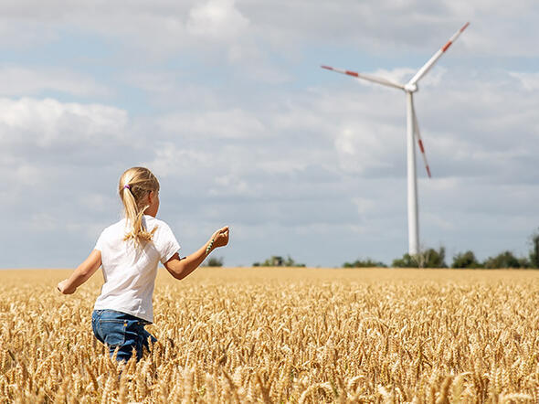 Junges Mädchen rennt durch ein Kornfeld mit einem Windrad am Horizont.